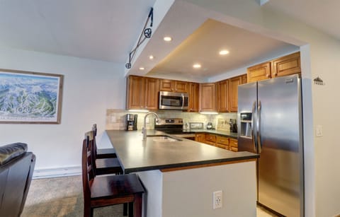 Modern kitchen with wooden cabinets, stainless steel appliances including a fridge and microwave, a black countertop with a sink, and bar seating. Wall art decorates the space.