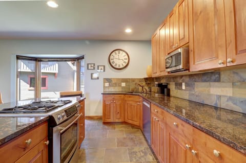 A modern kitchen with wooden cabinets, stainless steel appliances, granite countertops, a tiled backsplash, and a large wall clock above the sink. A bay window overlooks the exterior.