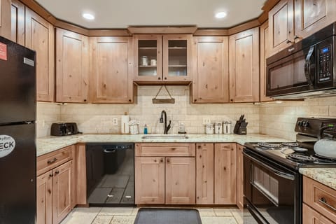 A kitchen with wooden cabinets, granite countertops, a black dishwasher, refrigerator, microwave, and stove. The sink is centered under cabinets with glass doors. Illuminated by ceiling lights.