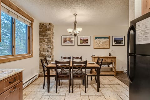 A dining area features a wooden table with six chairs, a chandelier above, wall art, a stone column, and a window. Part of a kitchen with a black refrigerator and granite countertops is visible.