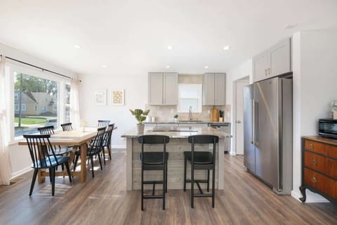 Open-concept kitchen & dining: sleek island with two bar stools faces a wood table for six by bright windows. Gray cabinets, stainless fridge, and wood floors create a harmonious flow throughout the space. ️✨