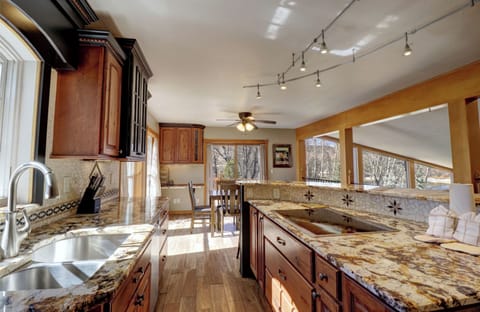 A modern kitchen with granite countertops, wooden cabinets, a double sink, and overhead track lighting. A ceiling fan is visible in the dining area beyond the kitchen.