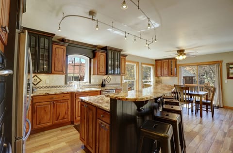 A modern kitchen with wooden cabinets, marble countertops, and bar seating. The space includes track lighting, a ceiling fan, and a dining area with a table and chairs near large windows.