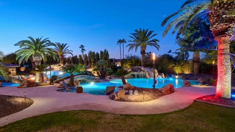 Resort-style pool with waterfalls, palm trees, and glowing torches at night.