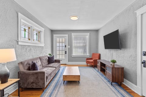 The living room features a gray tufted sofa, rust-colored accent chair, wood coffee table, wall-mounted TV, media console, area rug, decorative windows, and glass-paneled entry door.