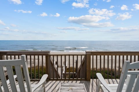 White deck chairs overlook a wooden railing, beach walkway, sandy shore, gentle Atlantic waves, and blue sky, perfect for an Oak Island, NC oceanfront vacation rental.