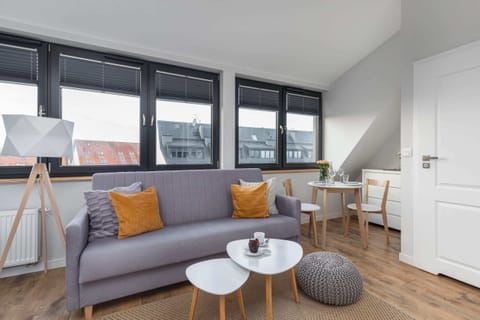 A bright and modern living room with a large gray sofa, decorative pillows in mustard yellow, and a round white coffee table. Natural light enters through large windows.