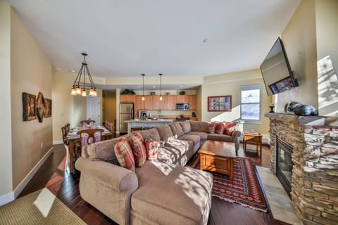 A modern open-concept living space featuring a brown sectional sofa, stone fireplace, wall-mounted TV, a dining area with a wooden table, and a kitchen with a breakfast bar and pendant lights.