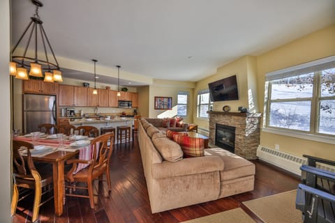 A cozy living and dining area with a large sectional sofa, a stone fireplace, and a mounted TV. The dining table is set for six. The open kitchen features wooden cabinets and stainless steel appliances.