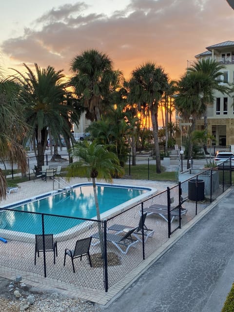 Swimming pool area with palm trees and lounge chairs against a dramatic sunset sky.