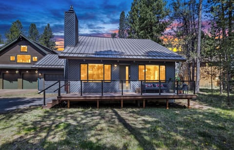 Modern mountain retreat surrounded by towering pines with a striking sunset backdrop. The property's sleek dark siding and warm interior lighting create a welcoming contrast to the natural landscape.