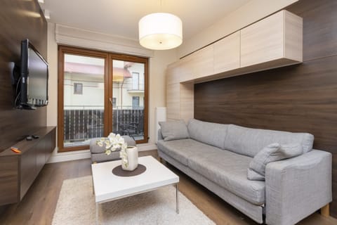 The living room features a modern gray sofa, a coffee table, and large windows that let in natural light. The space is cozy yet contemporary, with neutral tones and wooden flooring.
