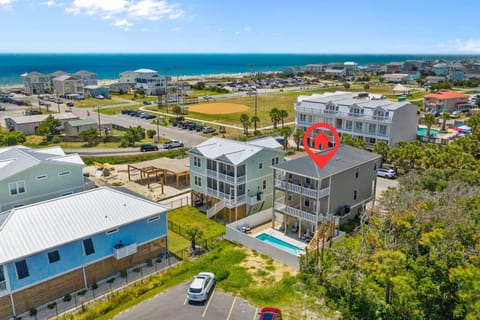 Drone view of marked coastal home with private pool, balconies, nearby park, beach access, ocean, homes, parking, and bright Oak Island, NC shoreline.