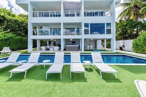Three-story waterfront villa with a private swimming pool, sun loungers, landscaped lawn, covered outdoor living and dining areas, expansive balconies, floor-to-ceiling windows, tropical greenery, and direct waterfront views.