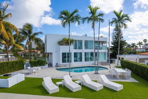 Contemporary waterfront estate featuring a private swimming pool, expansive pool deck, outdoor lounge seating, mature palm trees, manicured landscaping, floor-to-ceiling windows, and modern architecture with direct waterfront access and open views.