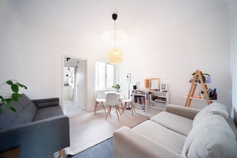 Bright and lively living room featuring two sofas, a dining table with four chairs, a floor lamp, and a mirror. The dining area is positioned near the window. The floor lamp illuminates a cosy corner, perfect for relaxation or reading.
