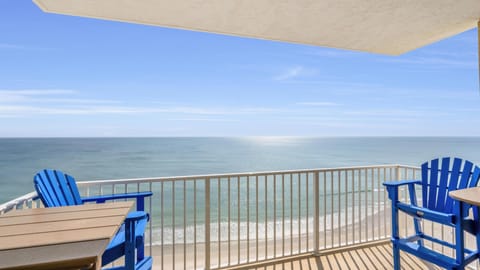 Balcony with blue Adirondack chairs and tan wood tables overlooks sandy beach and shimmering ocean.
