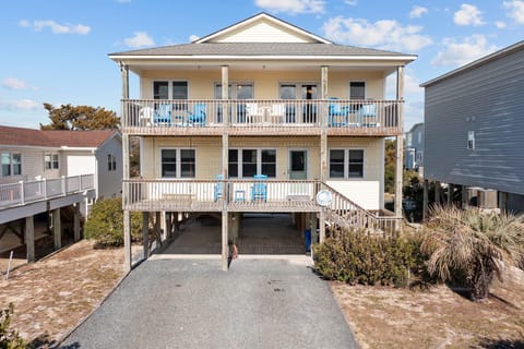 Elevated coastal home with two covered decks, outdoor seating, wood railings, stairs, covered parking, gravel driveway, shrubs, and blue sky.