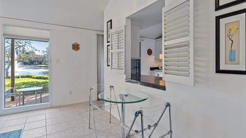 A bright room with tiled floor features a glass table with a chair, artwork on white walls, and a view of a lake with a fountain through a large window. An open shutter window connects to a kitchen area.
