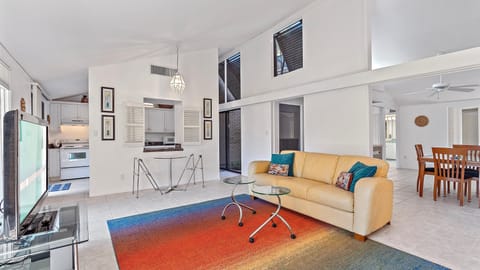 A modern living room with white walls, yellow sofa, colorful rug, glass coffee tables, and TV. Adjacent is a kitchen with white cabinets and dining area with wooden table and chairs.