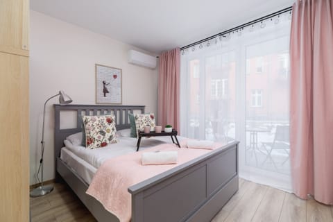 A bright bedroom with a double bed, soft pink bedding, and floor-to-ceiling windows.
