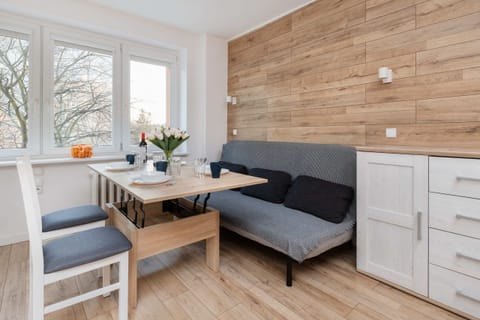 A cozy dining area with a wooden table, gray cushioned chairs, and a corner sofa, featuring natural light from large windows.

