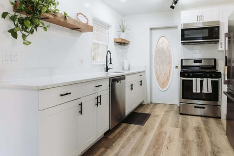 Prepare your favorite meals in this bright, airy kitchen featuring crisp white cabinetry, stainless steel appliances, and elegant quartz countertops. The open shelving with lush greenery adds a touch of warmth to this high-end, functional space.