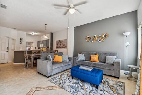 Welcome to a stylish living area, where soft gray couches and vibrant yellow pillows invite you to relax. A striking blue ottoman adds a pop of color, and natural light pours in through large windows, creating a warm atmosphere.