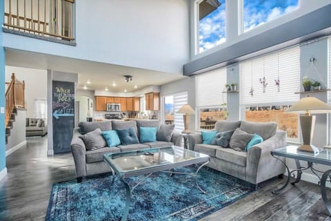 Double-height ceilings and large windows fill this open living area with natural light. Comfortable sofas frame a glass coffee table and blue rug, creating an inviting spot to gather, chat, or unwind while the kitchen and loft overlook the space.