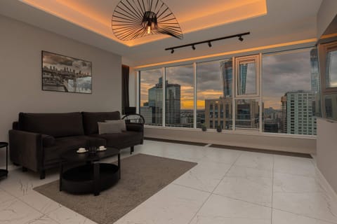 A spacious living room with a sleek, modern design, featuring a large sectional sofa, glass coffee table, and expansive windows that offer a stunning cityscape view, illuminated by warm ambient lighting from the ceiling.