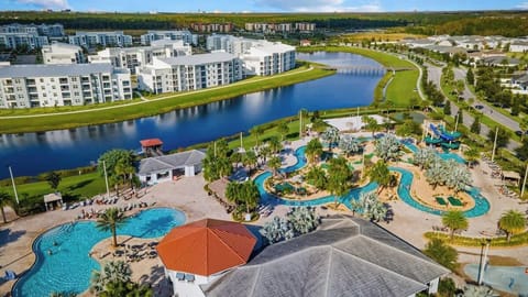 Stunning aerial view of a resort featuring a winding lazy river, large pools, and lush landscaping. Surrounded by modern white buildings and a serene lake, this vibrant setting offers a perfect mix of relaxation and fun for all ages.