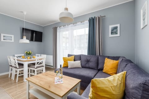 A cozy living room with a gray sofa adorned with colorful cushions. A coffee table and a small dining area in a bright, modern setting complete the space.