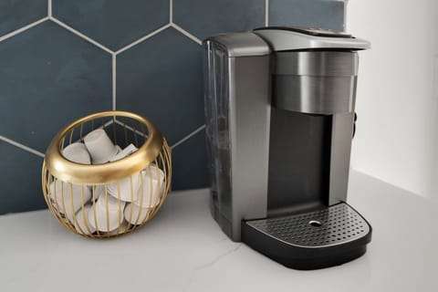 A sleek and modern kitchen corner with a single-serve coffee machine and a gold wire basket holding coffee pods, set against a marble countertop and a dark blue hexagonal tile backsplash.