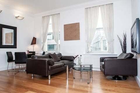 The spacious, bright living room with high ceilings creates an open, airy atmosphere. It's furnished with a comfortable sofa and armchair, perfect for lounging, and a sleek glass round table with chairs, providing a stylish and functional space.