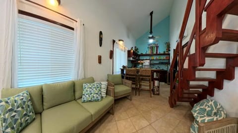 Green-cushioned sofa and armchair face a rustic wooden staircase beside a tiled floor and open kitchen bar.