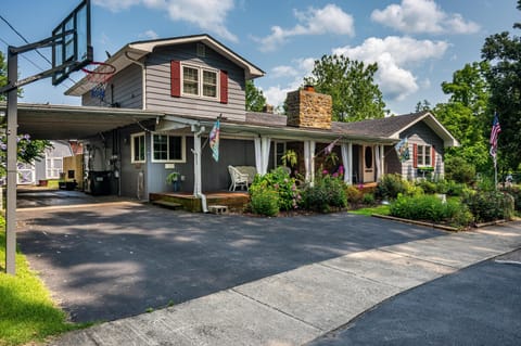 Gray siding with red shutters and a stone chimney frame the front porch with wicker chairs and garden beds.