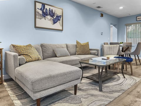 Open living room with sectional sofa, coffee table, abstract artwork, and dining area in background