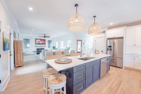Two round counter stools with woven seats are slipped under the left end of the kitchen island for extra seating. Stainless steel appliances and a navy kitchen island open up to the dinging area and living space in the background.