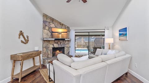 Living room with a stone fireplace, TV mounted above, white sectional sofa, wooden console table, wall decorations, and large sliding glass doors opening to a view of a swimming pool outside.