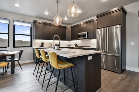 This modern kitchen boasts a spacious island with chic mustard-yellow barstools, ideal for casual meals or entertaining. Sleek cabinetry, stainless appliances, and airy windows elevate the space, blending style and function in an open-concept layout.