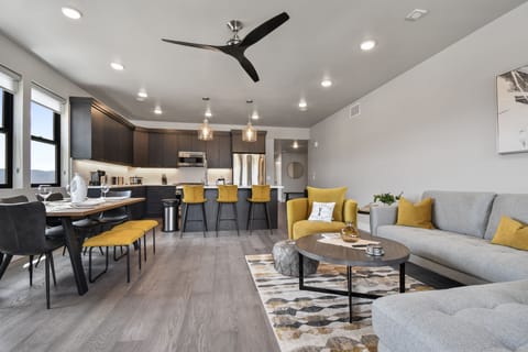 This open-concept living room flows seamlessly into the modern kitchen, featuring sleek cabinetry, a large island, and stylish pendant lights. Mustard-yellow accents add warmth, while natural light and wood floors enhance the airy, inviting vibe.