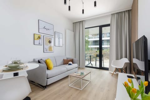 A modern living room with a sleek grey sofa, a coffee table, and a large flat-screen TV. The room is bright, with natural light coming in through floor-to-ceiling windows.