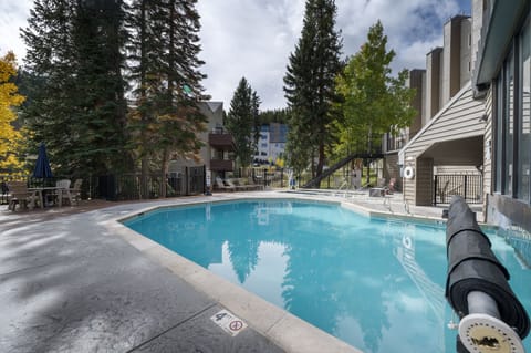 Outdoor swimming pool surrounded by trees and buildings, with lounge chairs and a lifebuoy visible. The pool is situated in a mountainous area, indicated by the surrounding landscape.
