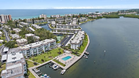 Aerial view of a coastal residential area featuring multiple apartment buildings, a swimming pool, docks with boats, a waterway, and the ocean in the background.