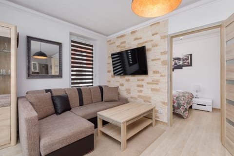 A cozy living room featuring a grey sofa with a wooden coffee table, a flat-screen TV mounted on the wall, and decorative wall panels with a stone-like design.