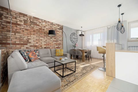 A modern living room with an exposed brick wall, a gray sofa, and a wooden coffee table. Industrial-style lighting and light wood flooring give the space a warm, urban feel.