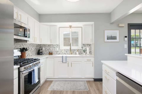 - Modern kitchen with stainless steel appliances and gas range   - White cabinets with gold handles and geometric tile backsplash   - Bright space with window above sink for a home style feel  