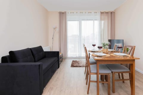 A bright and open living space with a cozy black sofa, a dining table for four, and large windows allowing natural light.