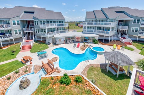Beachside resort featuring multi-level vacation rental buildings surrounding a curved swimming pool with spa, gazebo, and colorful outdoor amenities.