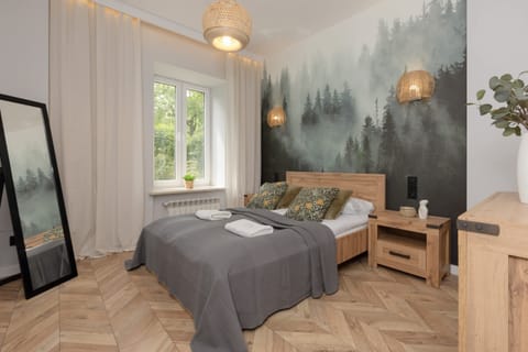 A cozy bedroom with a forest-themed mural, a double bed, soft lighting, and large windows for natural light.
